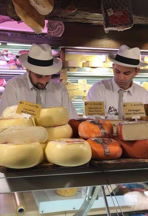 Two men wearing white hats examine various types of cheese