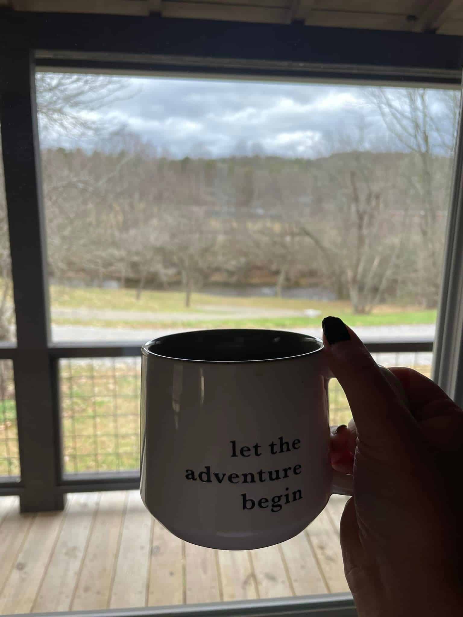 A hand gripping a mug inscribed with "Let the adventure begin,"