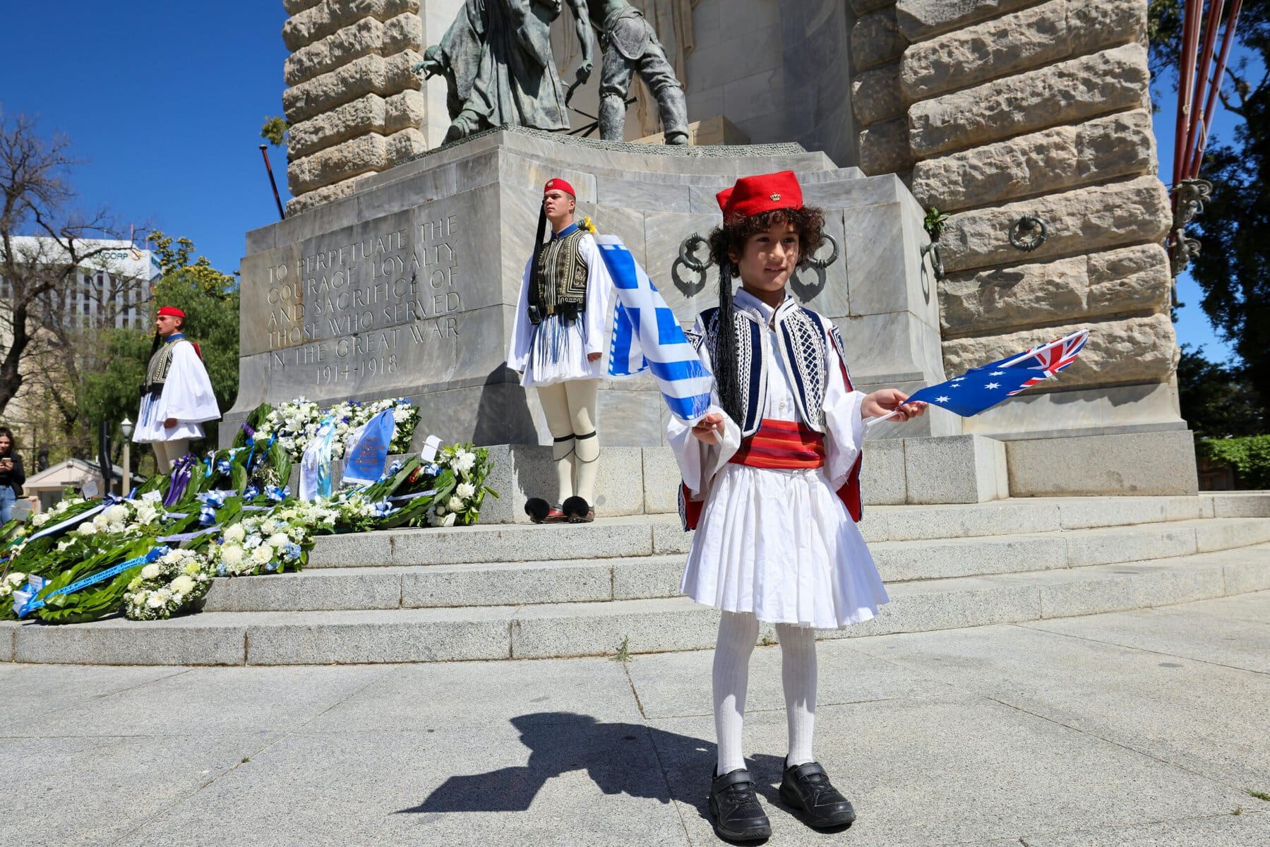 A young girl in traditional Greek attire poses in front of a historic monument