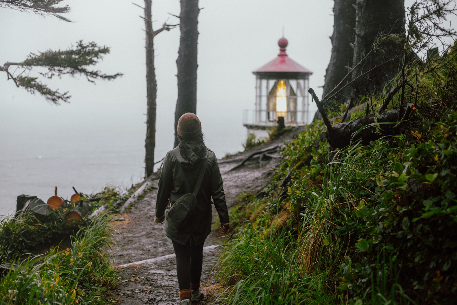 A lone hiker walks a foggy trail towards a coastal lighthouse in Oregon.