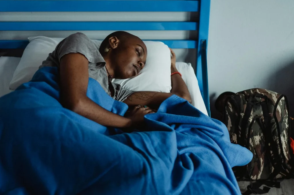 A woman rests peacefully in a hostel bed with a blue blanket and a backpack nearby, conveying travel fatigue.