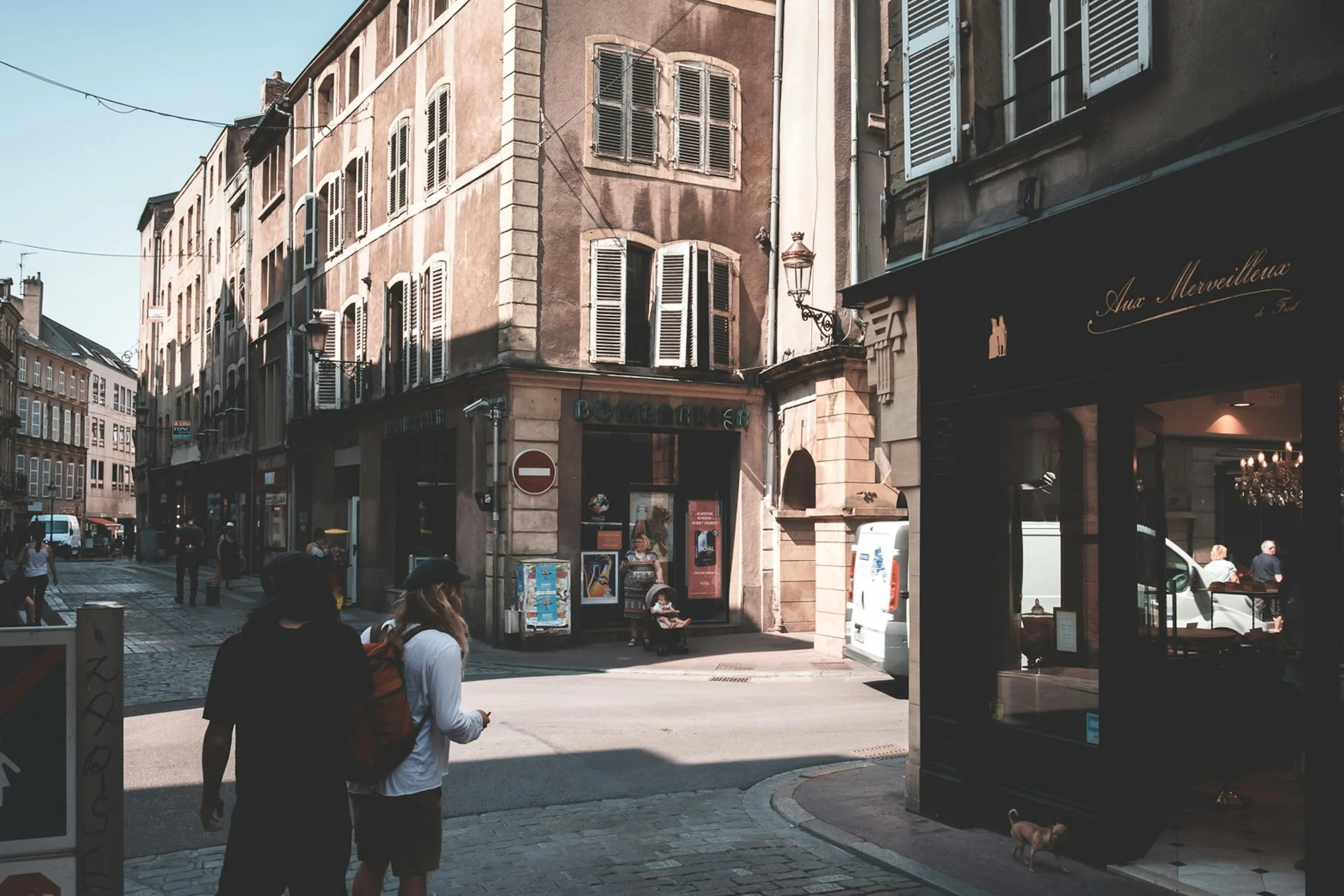 Back view of anonymous travelers with backpack strolling on paved walkway between aged buildings with stores on street in shadow.