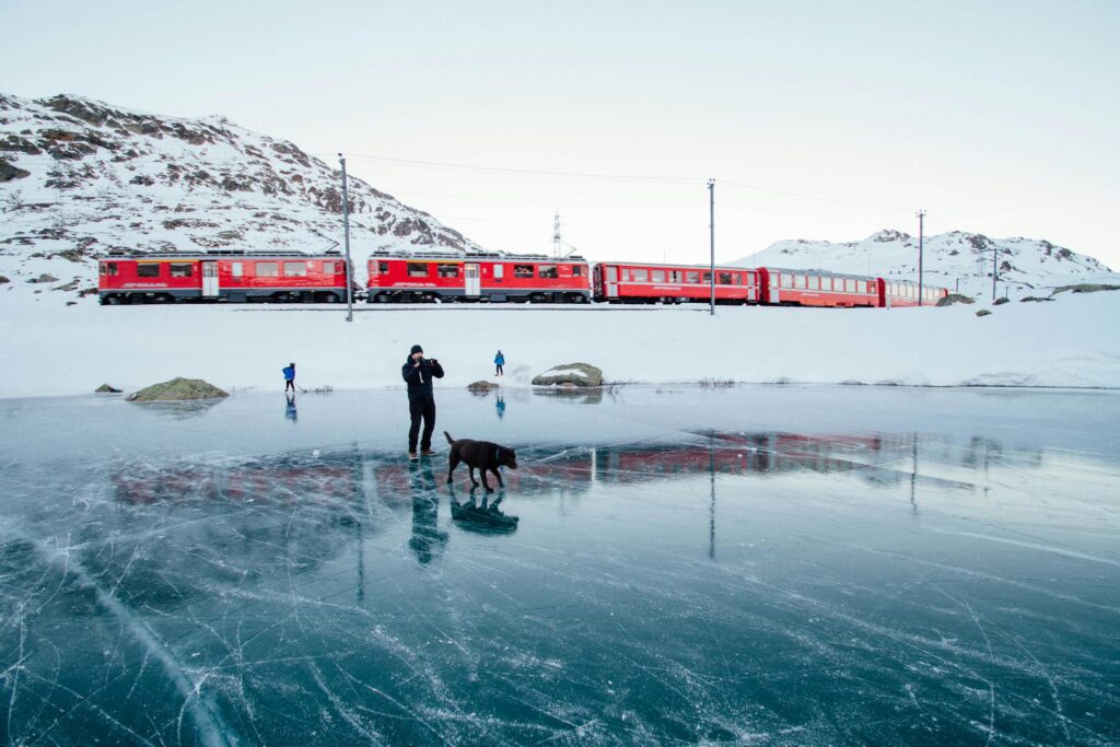 Capture of a train passing a frozen lake in Swiss Alps with people and a dog.