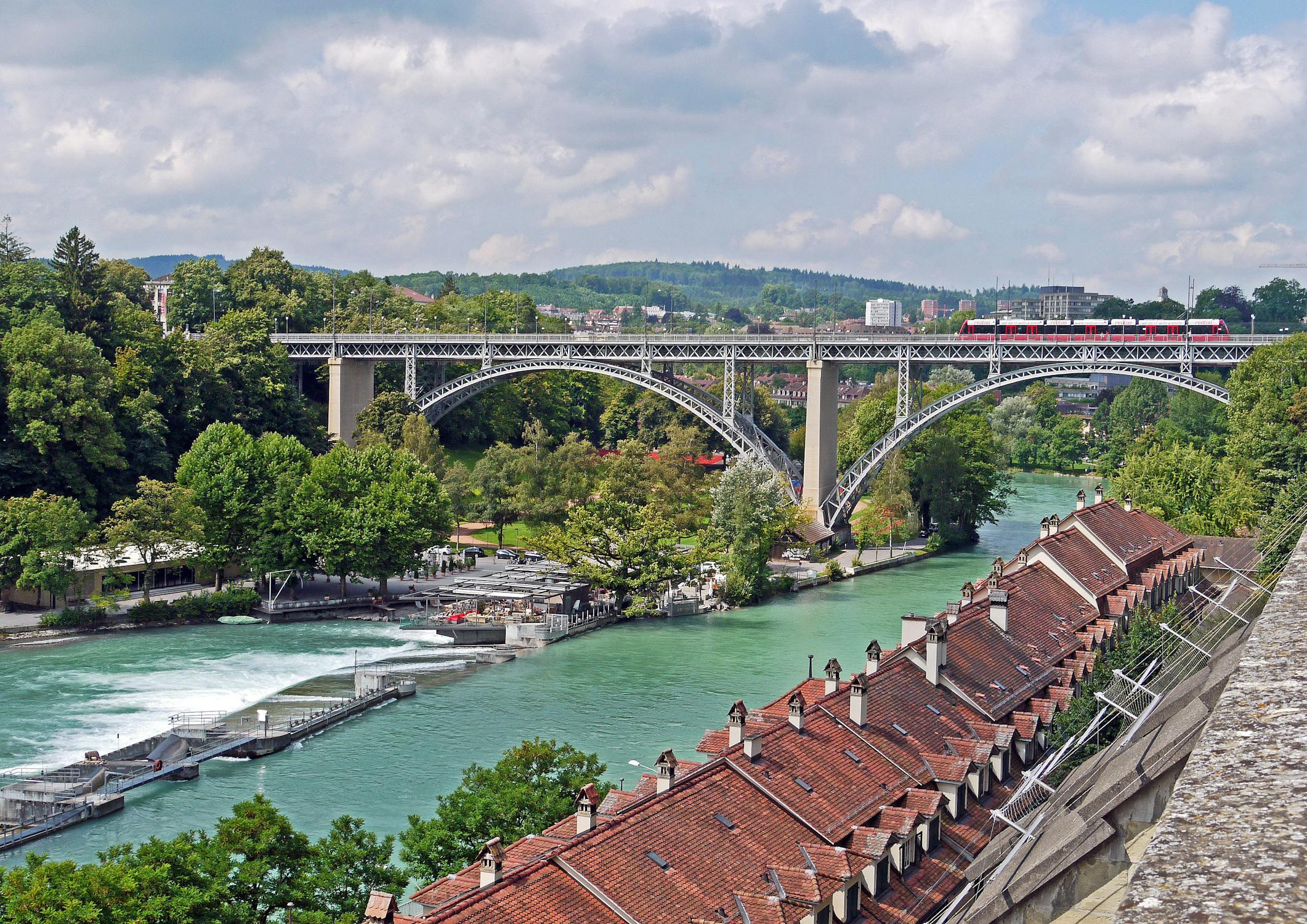 Panoramic view of the historic Kirchenfeld Bridge over the Aare River in Bern, surrounded by lush greenery and traditional rooftops.