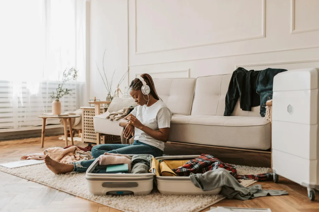 Young woman sits at home packing a suitcase while listening to music, surrounded by clothing items.